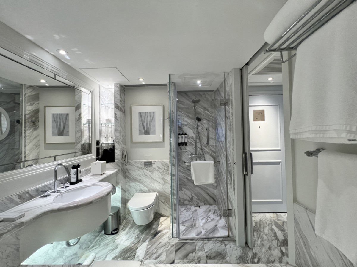 De badkamer Deze Deluxe kamer in The Langham heeft een prettig oppervlak. Je vindt hier een bad, wastafel, toilet en douche. Gemakkelijk kun je je hierin bewegen, echter vind ik de douche wel wat krap. De douche heeft verder een fijne handdouche en regendouche. De waterdruk is goed waardoor er een fijne waterstraal uit de regendouche komt. Ook de temperatuur is constant. Shampoo, zeep en conditioner zijn van het merk 34 Boulevard Saint Germain. Bij het bad is badzout en een blok zeep te vinden. Bij de wastafel zijn ook amenities van het merk beschikbaar, maar ook tandenborstels, oorsticks en een haarnetje. Onder de wastafel staat een weegschaal. Tussen de badkamer en slaapkamer is een raam. Deze is te blinderen met houten shutters. De badkamer is prettig in gebruik, netjes en schoon. Handdoeken zijn groot en zacht. Er is voldoende mogelijkheid aanwezig om deze op te hangen. 